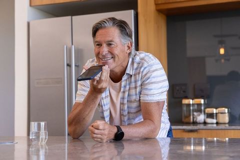Mature man speaking to smartphone on kitchen island, casual tech lifestyle, smiling