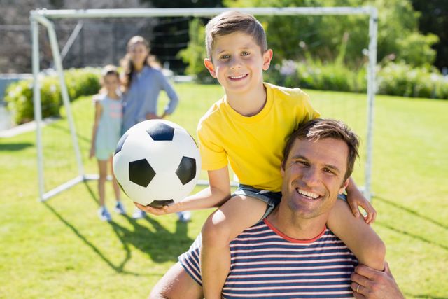 Father carrying his son on shoulders while holding a soccer ball in a sunny park. Family in the background near a goalpost. Ideal for concepts of family bonding, outdoor activities, parenting, and summer fun.