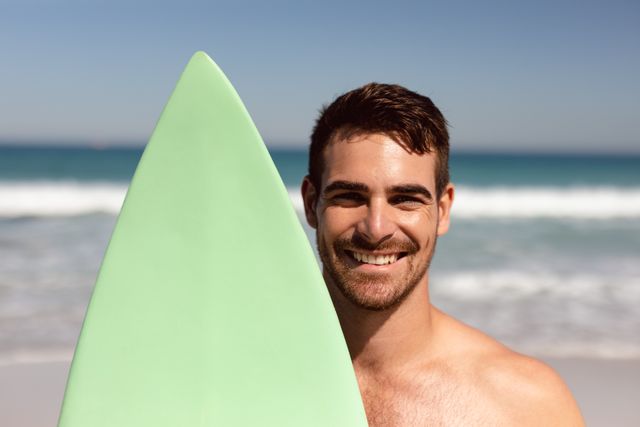 Front view of shirtless Caucasian young man with surfboard looking at camera on beach in the sunshine