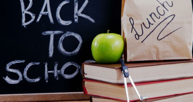 A stack of books with a green apple on top sits in front of a chalkboard featuring the words Back to School and a lunch bag to the side, with copy space. It symbolizes the start of a new academic year and the preparation involved.