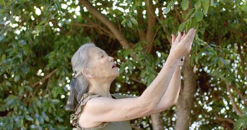 Senior asian woman reaching for tree branches in sunlit garden wearing smartwatch mindful pause