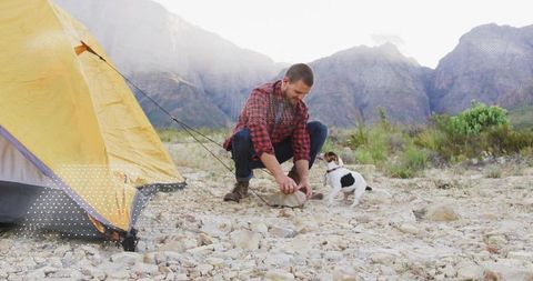 Man Securing Tent Near Mountainside with Dog