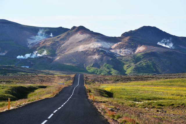Straight asphalt road stretches towards mountainous terrain with visible geothermal steam venting from the hills. Surrounded by lush, green fields, this remote and picturesque landscape is ideal for promoting travel and adventure advertisements, road trip inspirations and nature exploration guides.