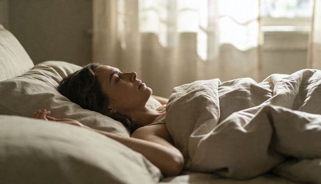 Woman reclining in sunlit bedroom, morning contemplation with neutral linen bedding