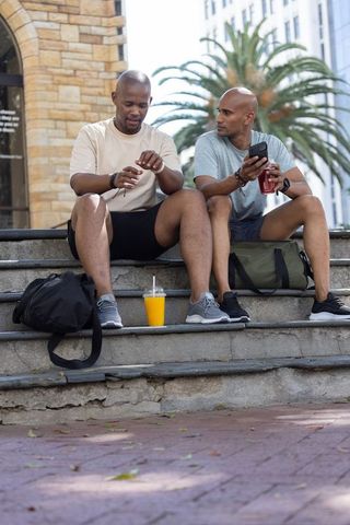 African american men checking smartwatch and using smartphone on urban plaza steps