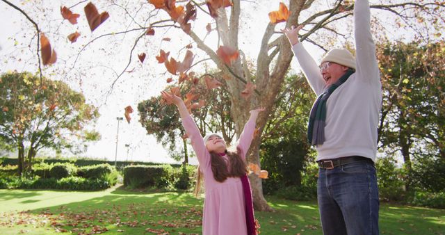 Father and daughter enjoying a playful moment with autumn leaves in a beautiful park. Both are wearing winter clothes and smiling while throwing leaves in the air. Ideal for seasonal greetings, family-oriented content, and promoting outdoor activities.