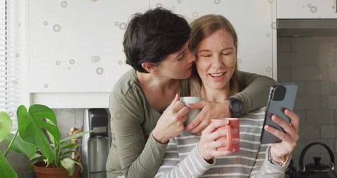 Affectionate female couple sharing coffee and scrolling smartphone in bright kitchen
