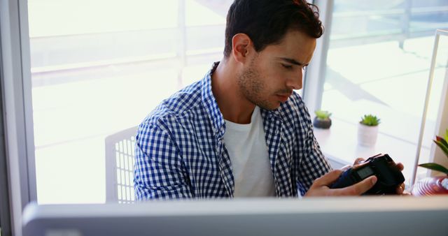 Young male photographer adjusting camera settings in modern office workspace. He is sitting at a desk, surrounded by decor and office plants, and wearing casual attire. This image is ideal for websites or promotional materials related to photography, office environments, digital workspaces, or career-focused content.