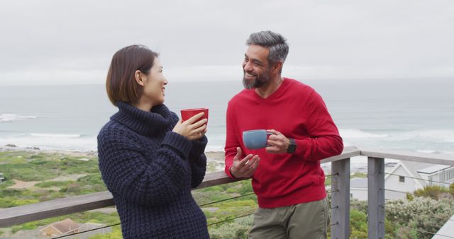 Middle-aged couple wearing sweaters chatting and smiling on a balcony with a beautiful ocean view. Both holding coffee mugs in a relaxed posture. This image can be used in lifestyle blogs, vacation brochures, and articles about relationships or outdoor living.
