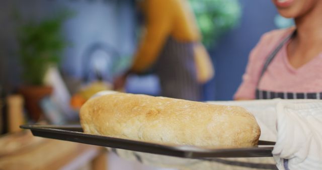 Person taking out freshly baked bread from oven while wearing apron and oven mitts in kitchen. Perfect for cooking blogs, bakeries, home cooking advertisements, and recipe illustrations.