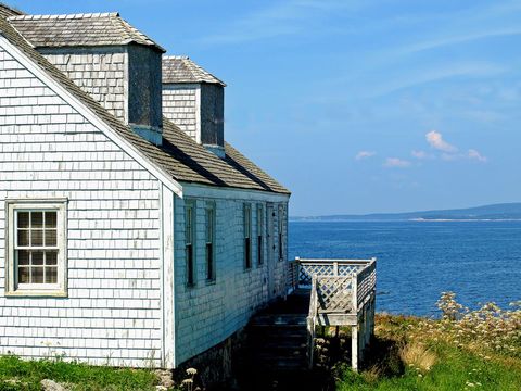 Weathered white shingle cottage on rugged coastline overlooking calm blue ocean