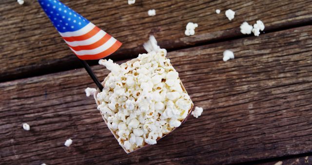 This image depicts a small portion of popcorn adorned with an American flag, set against a rustic wooden background. Perfect for use in content related to American national holidays like Independence Day or Memorial Day, food blogs highlighting festive snacks, or advertising for events and celebrations.