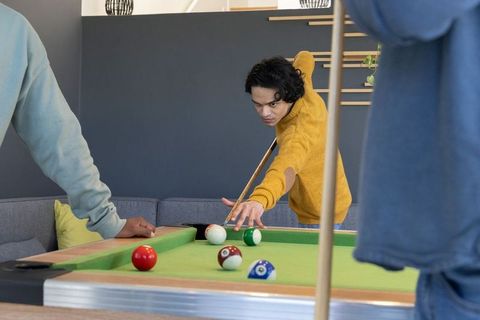 Asian man in mustard sweater aiming cue while playing pool with friends in modern lounge