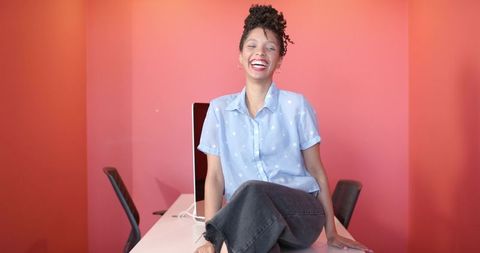 Young woman sitting on desk laughing in modern coral workspace with laptop and chairs