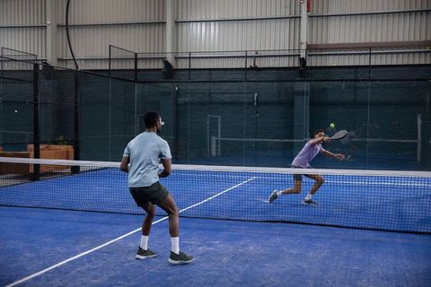 Diverse male opponents playing padel on indoor blue court lunging to return ball
