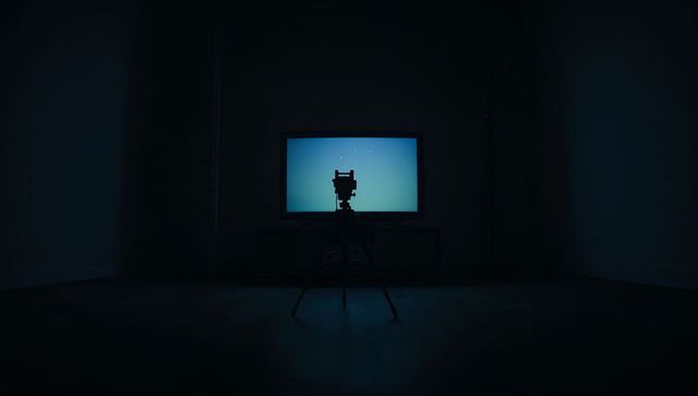 Silhouetting vintage camera on tripod facing cyan display in dark minimal studio