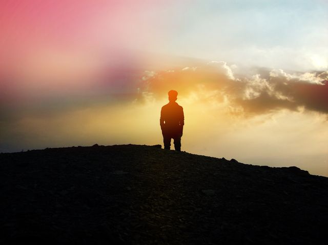 Silhouette image of a man standing on a hill during a vibrant sunset, with colorful sky and clouds in the background. Suitable for themes related to solitude, contemplation, inspiration, nature tranquility, and peacefulness. Ideal for website banners, motivational posters, and backgrounds.