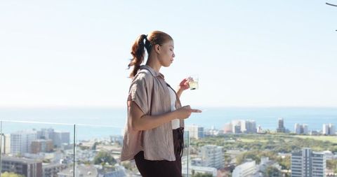 Young woman enjoying drink on rooftop balcony overlooking ocean and city skyline