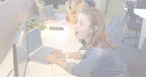Young woman leaning forward typing on laptop with headphones in modern open-plan office