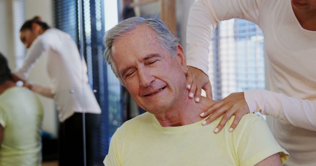 A senior Caucasian man receives a shoulder massage from a therapist, in a physical therapy or wellness setting, with copy space. His relaxed expression suggests relief and comfort during the therapeutic session.