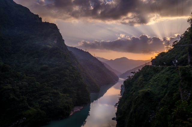 Sunlit river winding through lush green mountains, silhouetted by sunset sky with dramatic clouds and sunrays. Ideal for use in travel blogs, landscape photography collections, nature-focused publications, and inspirational social media posts.