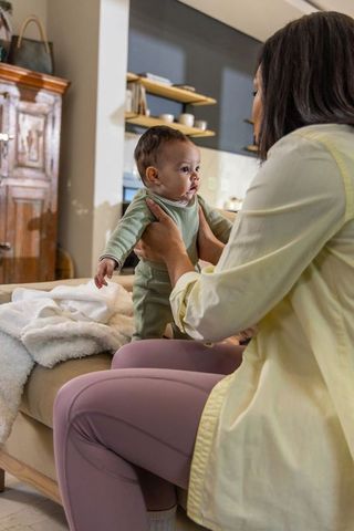Mid adult woman holding infant upright on ottoman supporting under arms in cozy home