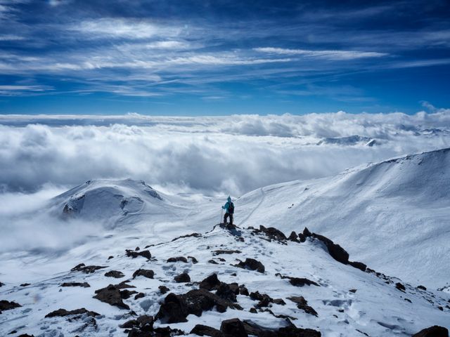 Single hiker standing on a snow-covered mountain summit with a panoramic view of clouds and sky. Perfect for illustrating themes of adventure, exploration, solitude, and the majesty of nature. Ideal for travel websites, adventure blogs, or inspirational posters.