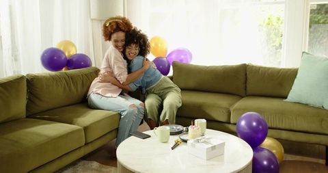 Two African American women hugging on olive sofa celebrating birthday with balloons