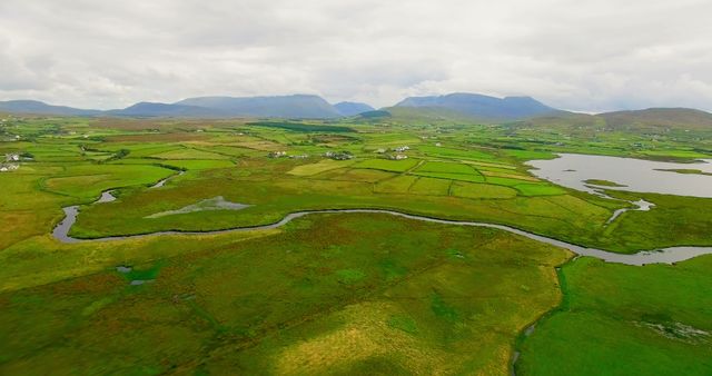 Aerial view showcases a lush, green landscape dotted with bodies of water and distant mountains under a cloudy sky. The patchwork of fields and waterways highlights the rural beauty and tranquility of the region.