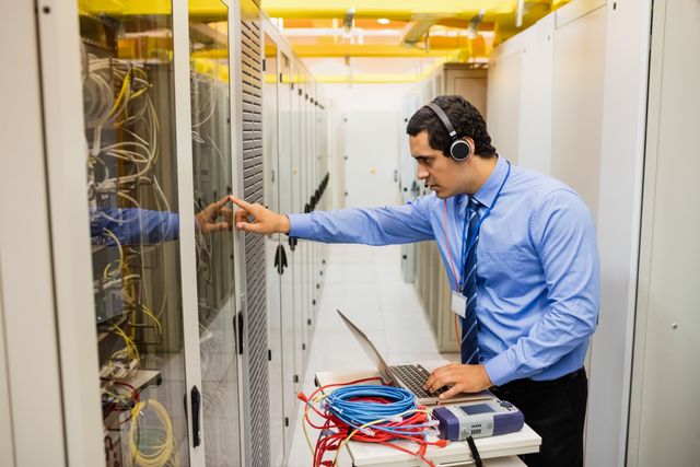 Technician in a server room monitoring systems using a laptop while wearing headphones. Network cables are visible on the table, indicating ongoing maintenance or troubleshooting. Ideal for illustrating IT support, data center operations, network management, and technology maintenance.