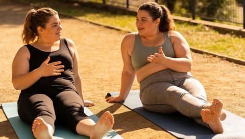Two women practicing yoga and chatting on mats in sunlit park for fitness and wellness