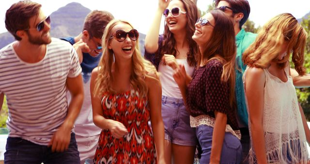 Young adults enjoying an outdoor party in warm weather. They are all wearing sunglasses and casual clothes, laughing and having fun against a backdrop of mountains. This image can be used for promoting social events, summer gatherings, outdoor activities, or young adult lifestyle products.