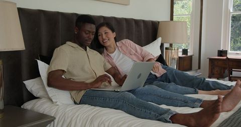 Diverse Couple Relaxing on Bed Using Laptop