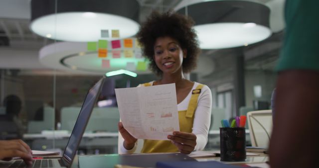 This image shows a young professional woman reading a document at her desk in a modern office. Her laptop is open, and colorful sticky notes are on a glass wall behind her, illustrating a collaborative and innovative work environment. This can be used for content related to business, teamwork, female professionals, corporate culture, workplace diversity, and modern offices.