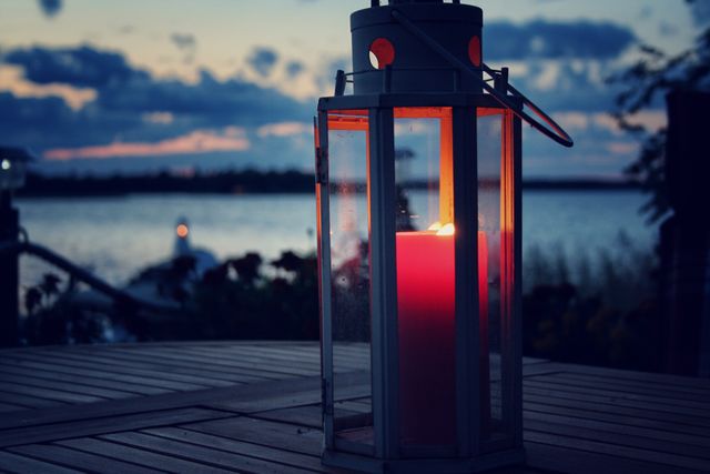 Lantern with a candle glowing softly in the evening by a serene lakeside. Perfect for illustrating outdoor tranquility, romance, or peaceful moments. Ideal for use in blogs, websites promoting outdoor activities or romantic getaways, and social media posts focusing on relaxation.