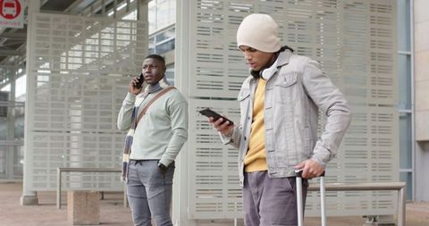 Two diverse male commuters checking smartphones at outdoor transit stop with luggage