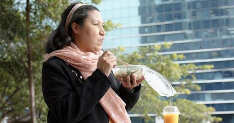 Mature woman enjoying quick urban lunch with salad and juice near office towers