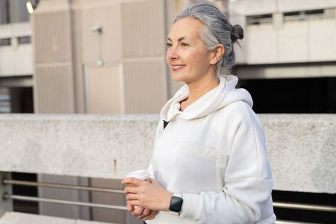 Mature woman wearing white hoodie holding coffee on urban rooftop, smiling and reflecting