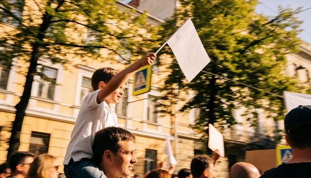 Child waving blank white flag on parent's shoulders during sunny urban protest parade crowd