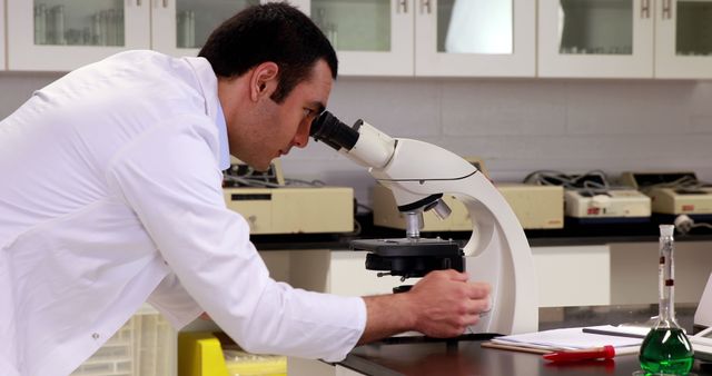 A scientist in a lab coat is examining samples under a microscope in a laboratory. He is focused on his work with various lab equipment and notes around him. This image can be used to represent scientific research, laboratory settings, scientific discoveries, educational materials, and medical research. It highlights precision, concentration, and the scientific method in action.