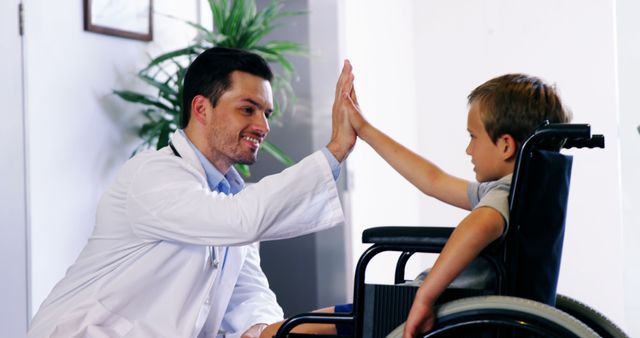Doctor high-fiving young patient in wheelchair, capturing positive interaction and support in medical care. Can be used for healthcare promotion, pediatric care, disability support, and hospital advertisement.