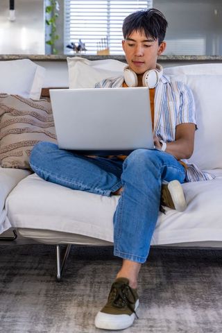 Asian teen studying on white sofa with laptop and headphones in cozy modern living space