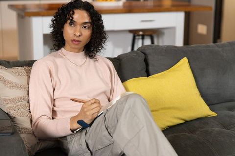 Non-binary person journaling on gray sofa in cozy open-plan home, focused and reflective