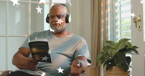 Senior man relaxing in sunlit living room reading endless sunsets while wearing headphones
