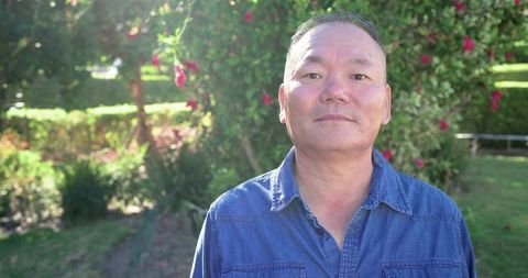 Senior asian man standing in sunlit garden wearing denim shirt