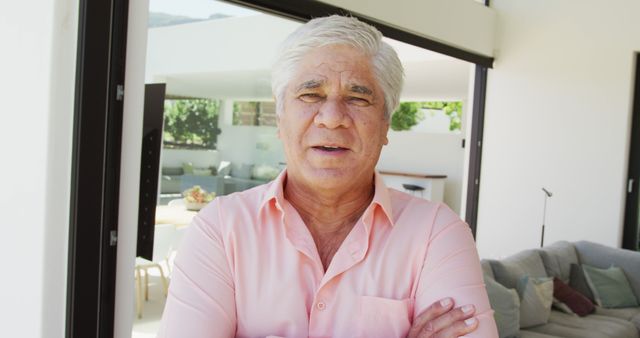 Mature senior man standing with crossed arms in a modern, contemporary living room with natural light. Ideal for uses in retirement ads, health and lifestyle promotions, senior living features, and home-related publications.