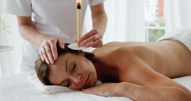 A woman is receiving an ear candling therapy at a spa, lying peacefully on a massage table with a candle lit in one ear. Perfect for illustrating wellness, self-care, and alternative therapy concepts. Useful for websites, blogs, or advertisements focused on holistic health, spa treatments, relaxation techniques, and natural healing practices.