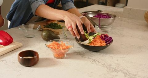 Female friends preparing colorful avocado and edamame buddha bowl on marble countertop