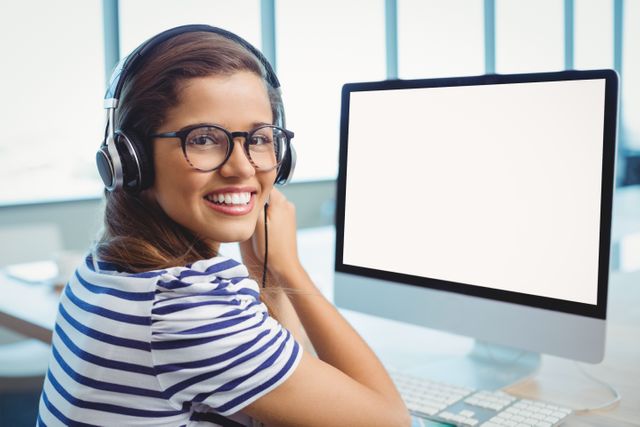 Young female graphic designer wearing headphones and glasses, working on a computer in a modern office. Ideal for illustrating concepts related to creative professions, digital design, modern work environments, and professional women in technology. Suitable for use in articles, blogs, and marketing materials focused on graphic design, workplace culture, and technology.