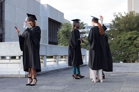 Multicultural female graduates celebrating on campus terrace holding diplomas and smartphones